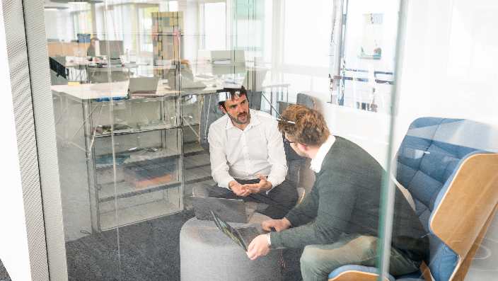 Two people are sitting in a room talking to each other. They are visible through a pane of glass.